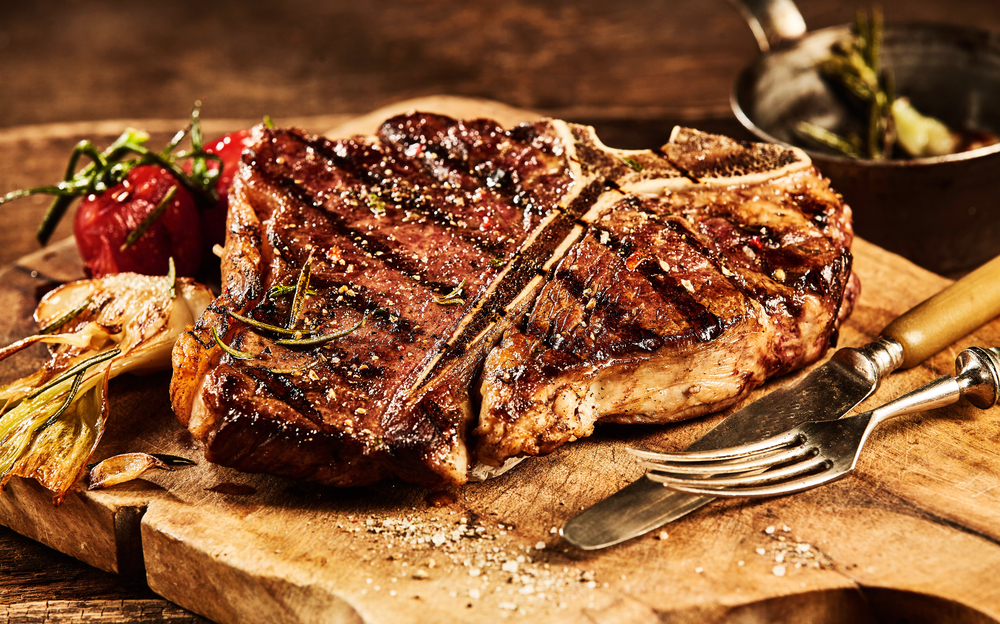 A large grilled steak on a board with cutlery and grilled tomatoes