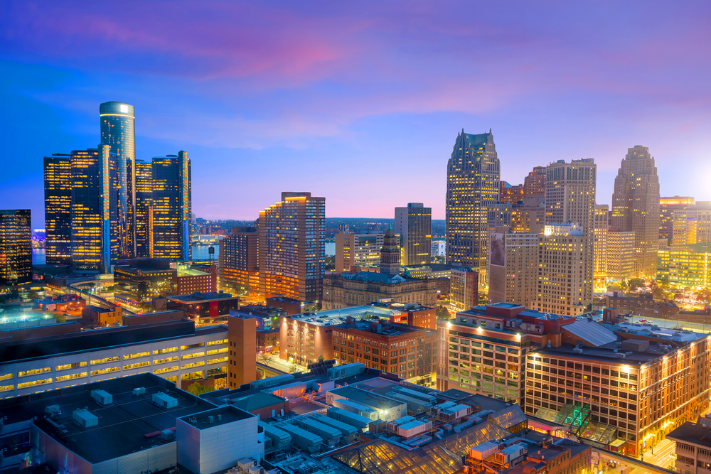 The Detroit skyline at twilight. The sky is blue, pink, and purple. The lights in the buildings are all on.