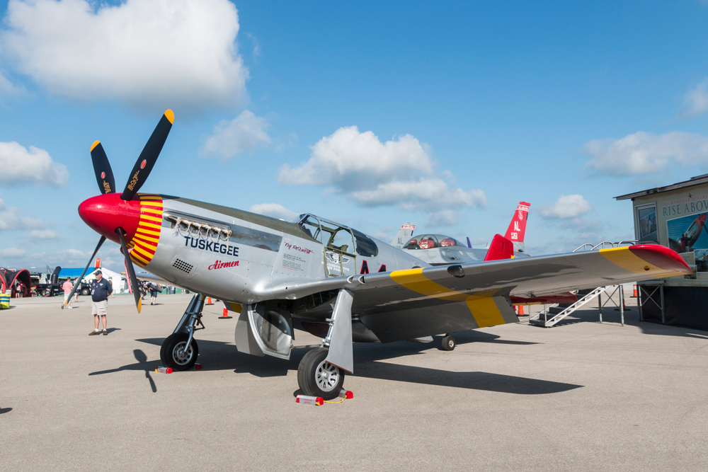 An airplane used by the Tuskegee Airmen in World War II on a tarmac. It is silver with red and yellow accents painted on it. 