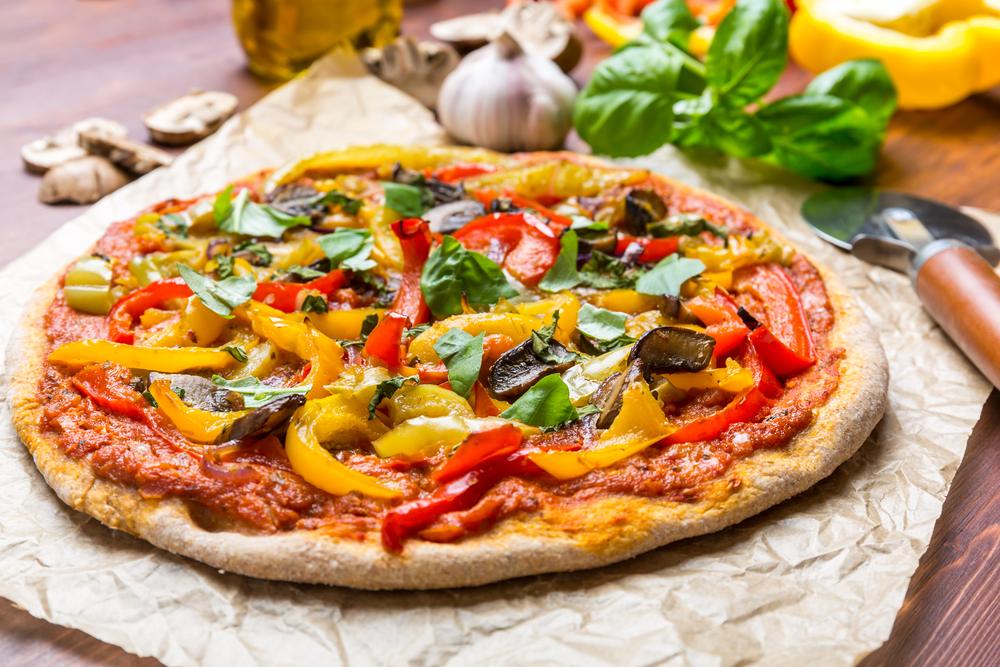 A large vegan pizza with peppers, mushrooms, red sauce, and fresh basil. It is on a crumpled piece of butcher paper. You can see a pizza cutter and other fresh vegetables around it.