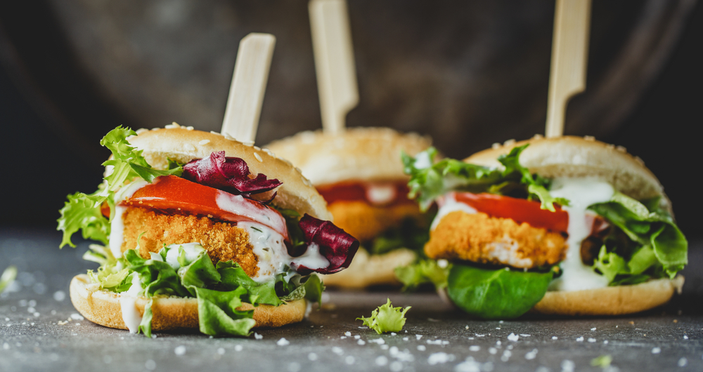 Vegan chicken sandwich sliders like ones you can find in restaurants in Detroit. They have lettuce, tomato, and a creamy sauce on them. They have toothpicks in the middle of them.