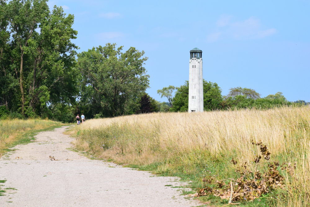 A view of the Art Deco William Livingston Memorial Lighthouse on Belle Isle. Around it is a field with tall grass, a dirt path, and a wooded area. 