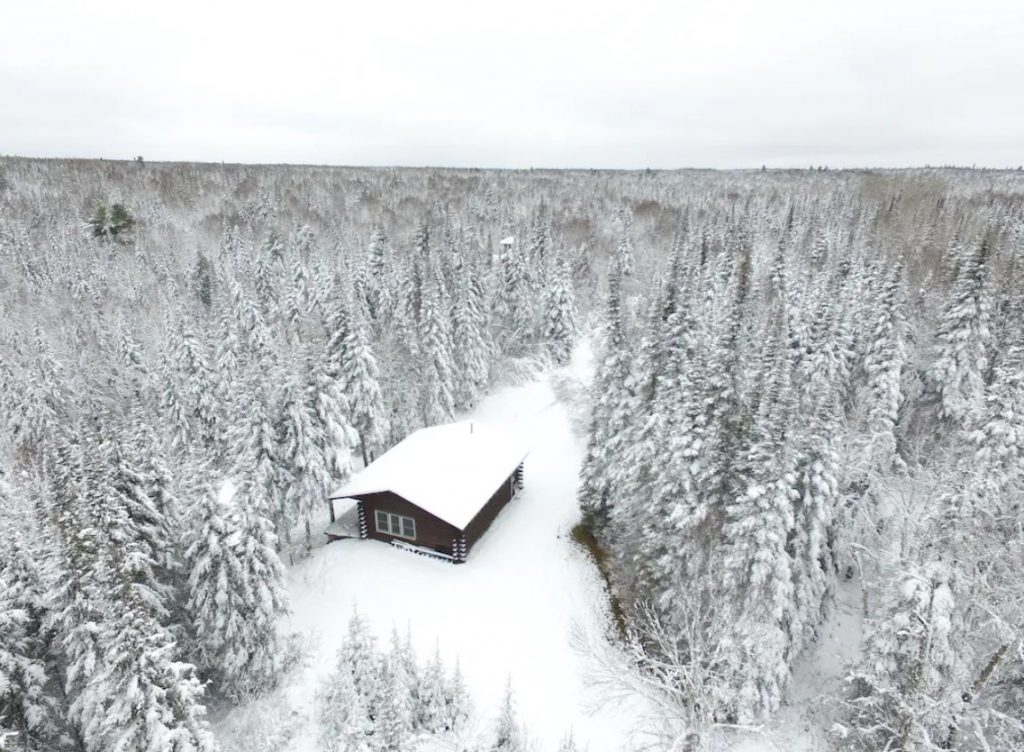 Wintery overhead view of cozy Minnesota cabin with snow on its roof and forested trees surrounding it all snow covered.