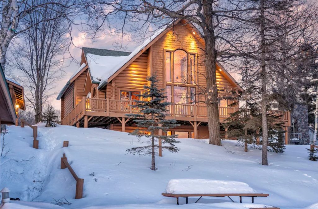 Large log cabins in Minnesota at sunset with snow on ground, illuminated by accent lights. Large windows everywhere.