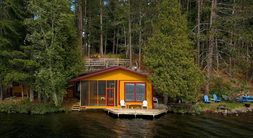 Yellow cabin in MN with red trim sits directly on lake waters surrounded by green forested trees.