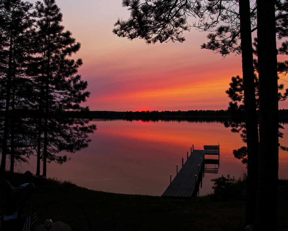 Orange and purple sunset sky with smooth lake and wooden pier in foreground at one of the cozy cabins in Minnesota.