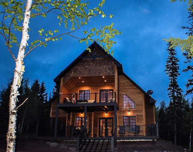 Wooden MN cabin illuminated by accent lights, with many windows, blue night sky in background.