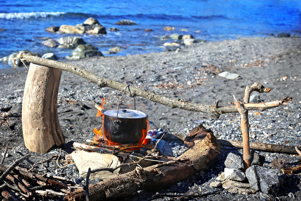 A makeshift firepit on a rocky lake shore. There is a black metal pot hanging over a fire from a large branch. 