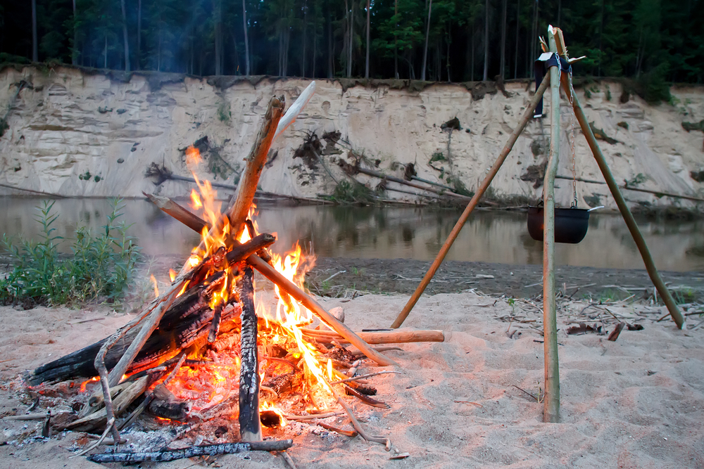A roaring bonfire on the shores of a lake. next to it is a pot on a tripod. In the background you can see the river and tall cliffs. 