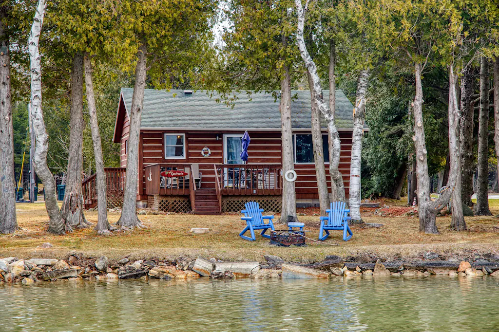 A small brown cabin with water in the foreground and two blue chairs around a fire pit.