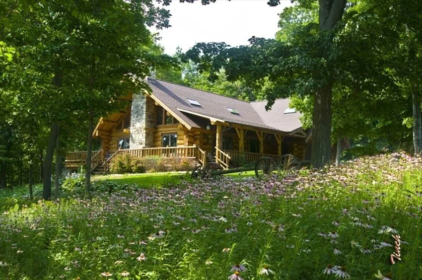 A large log cabin among trees with a wildflower field in the foreground