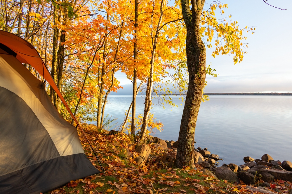 A tent on the shores of a large lake. It is fall and the leaves on the trees are yellow and orange. The sun is setting on the lake. 