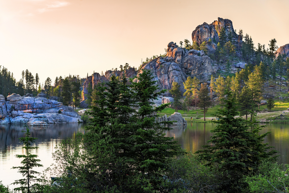 One of the amazing views at Custer State Park in South Dakota. There is a large mountain rock formation, a lake, grassy areas, tall trees, and more.