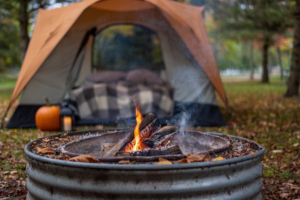 A bonfire at a campsite in Michigan in the fall. You can see a tent with a large raised bed with a quilt on it. Outside the tent is a pumpkin and a lantern. You can see dead leaves on the ground. 