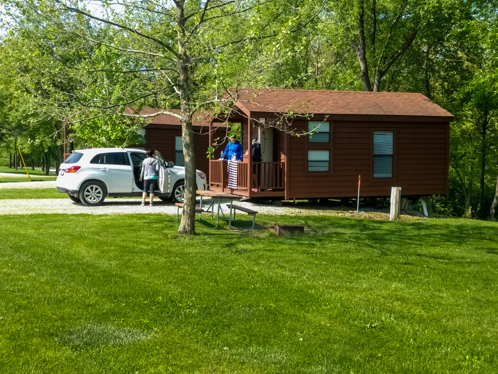 Two cabins in the woods with a lawn next to them. You can see a car in front of one and people standing around. One of the best places for camping in Indiana.