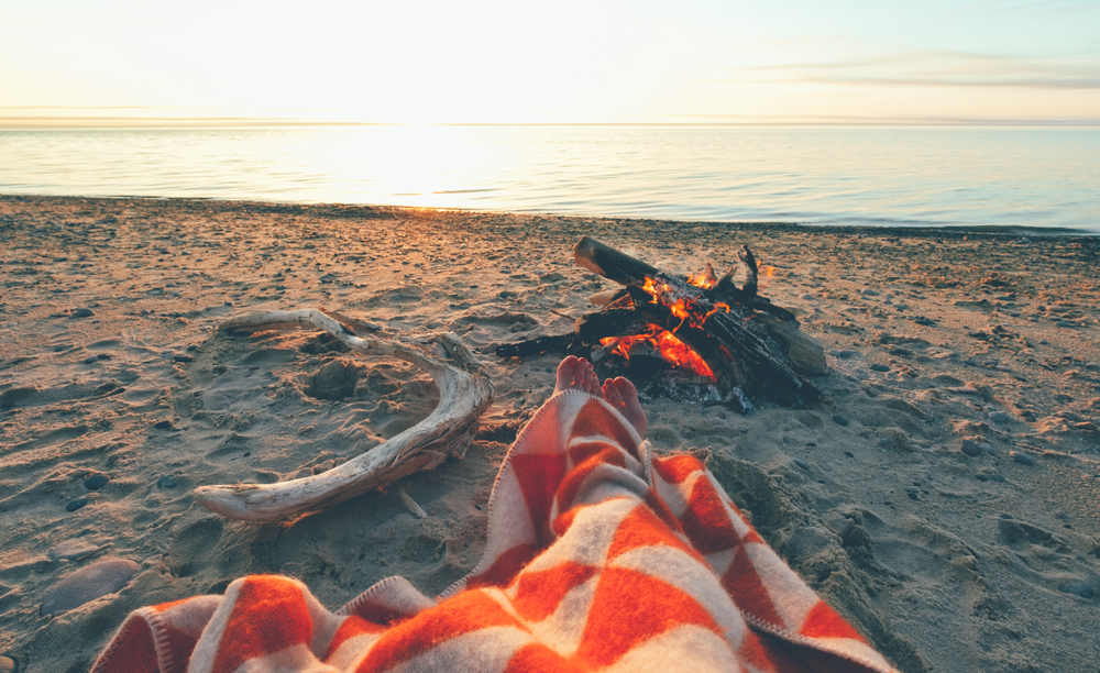 A person sitting on the shore of the lake with an orange and white blanket in their lap. Their feet are near a small bonfire on the shore. You can see the lake for miles. 