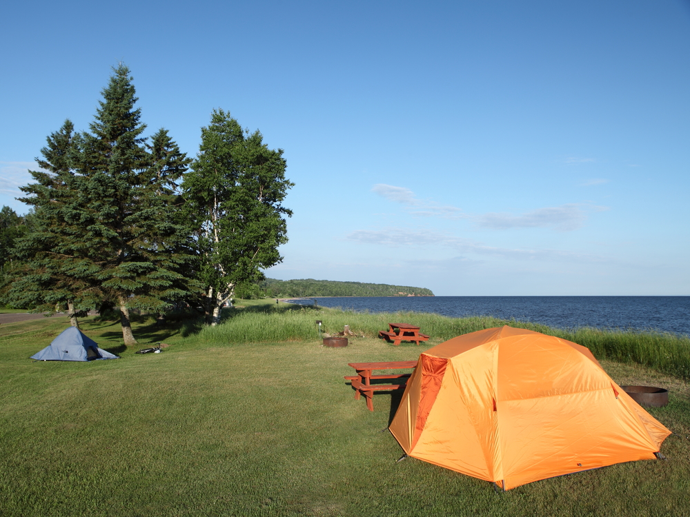 A large orange tent on the shore of a large lake. You can also see a smaller blue tent. The campsites have picnic tables and firepits. The campsites are grassy and there are some trees. 