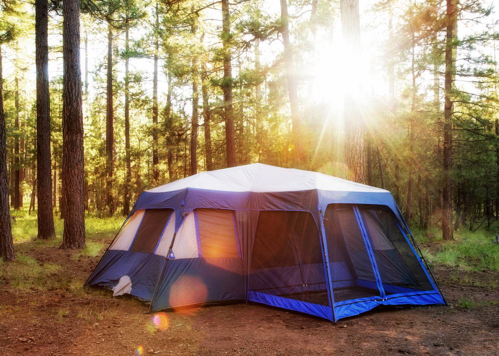 A large multiple chamber blue tent in the woods like you'll finding camping in Indiana. The sun is shinning through the trees onto the tent. 