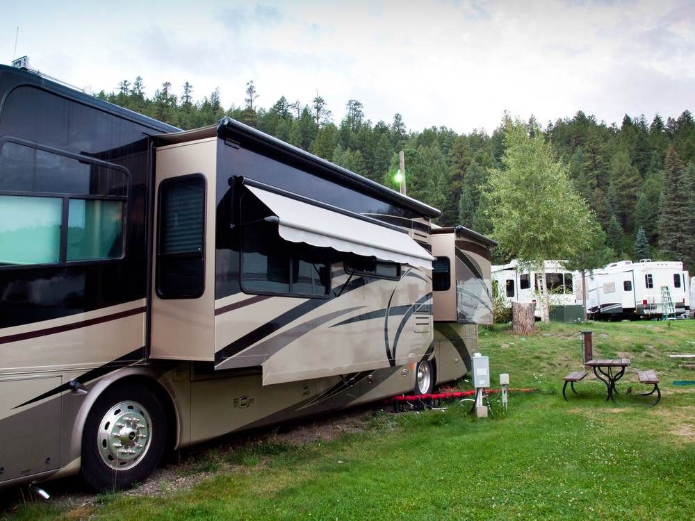 A large and luxurious RV that has several pop-outs at a camp site. In the background you can see other RVS and a forest of dense trees. 