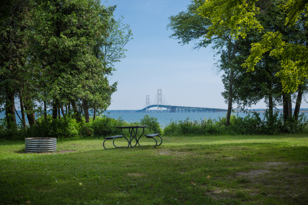 A campsite on the edge of the lake where you can see the Mackinaw Bridge in the distance. There is a firepit and a picnic table on the campsite. One of the prettiest places for camping in Michigan. 