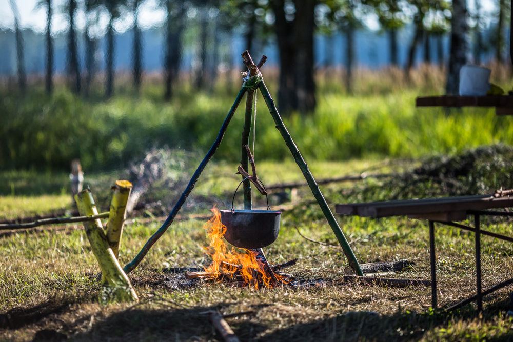 A primitive cooking set up over a fire at a secluded woodland campsite. There is a pot hanging over a fire using a pyramid-like structure made of branches. 