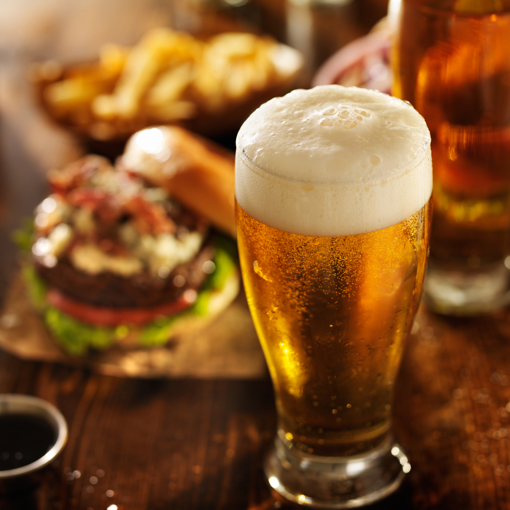 Beer on a table in foreground with a loaded burger in the background