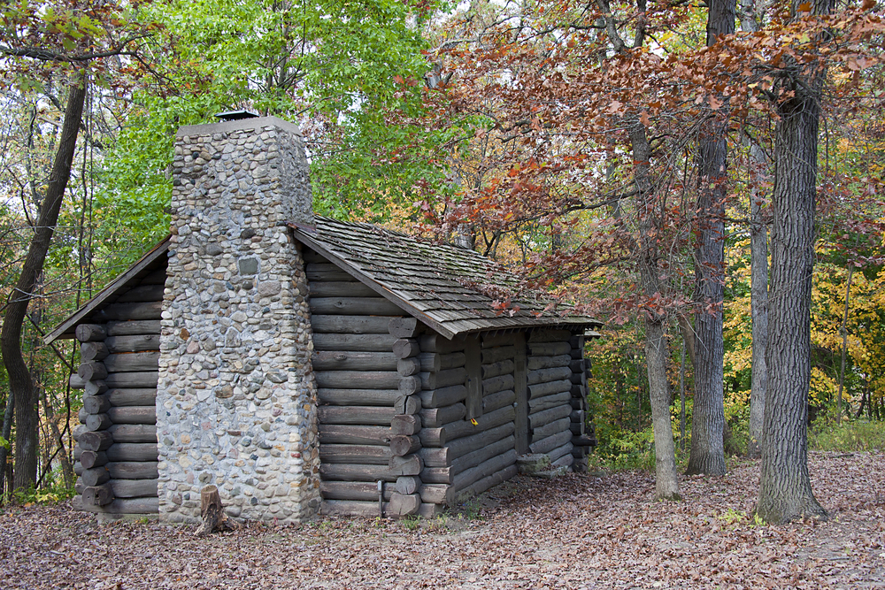 A rustic style cabin with a stone chimney. It is surrounded by trees and it is fall so there are dead leaves on the ground. Some of the trees have red and yellow leaves. 