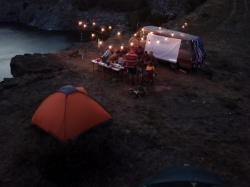 A group camping on the edge of a lake with an RV that has string lights over a picnic area and a small orange tent. It's a pretty area similar to other campsites in Indiana. A great place for camping in Indiana