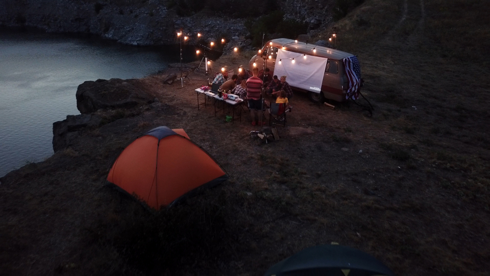 A group camping on the edge of a lake with an RV that has string lights over a picnic area and a small orange tent. It's a pretty area similar to other campsites in Indiana. 