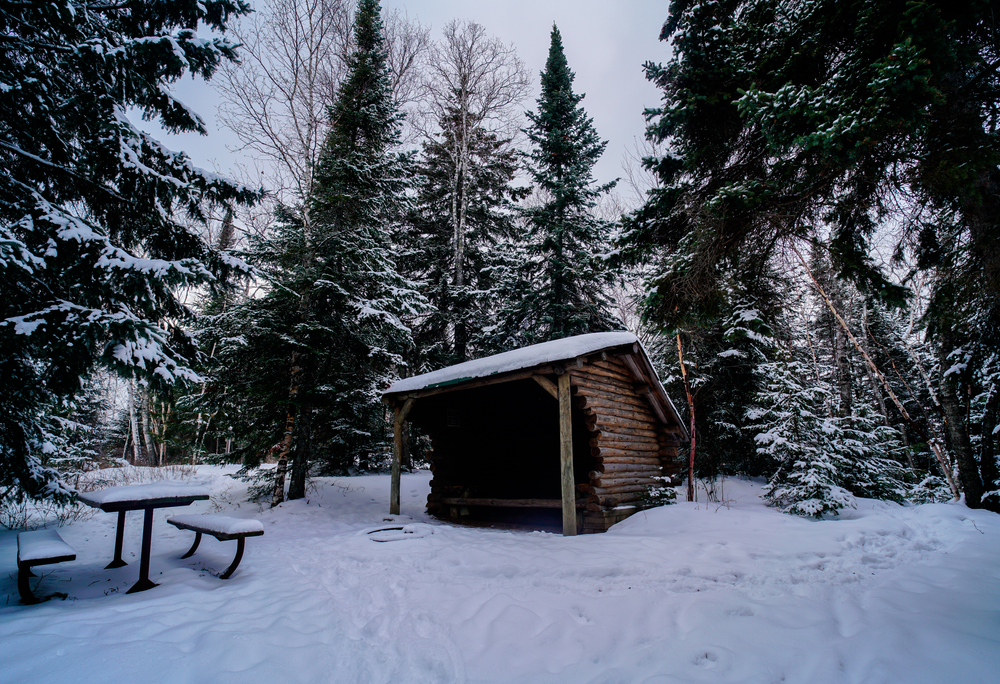 A small wooden log hut on a small campsite. You can see a picnic table and a firepit covered in snow. It is one of the best places to try camping in Indiana in the winter. 