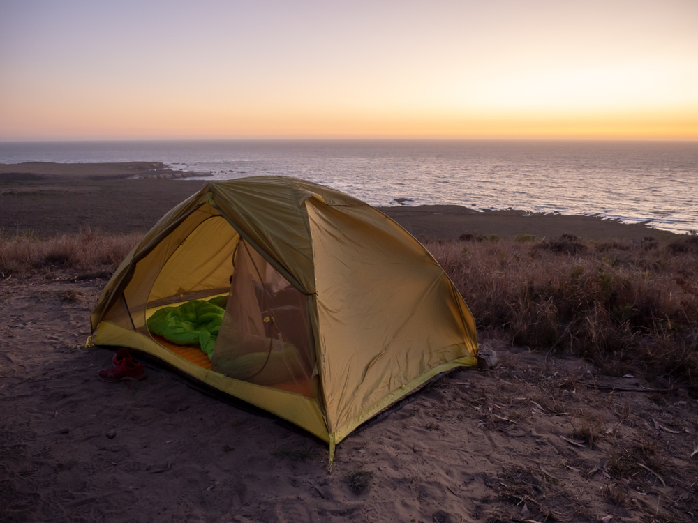 A small green tent on the shores of a large lake or ocean at sunset. It's similar to campsites you can use at one of the best camping in Indiana parks. 