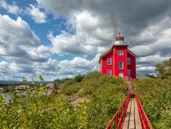 A red lighthouse at the end of a wooden boardwalk surrounded by greenery