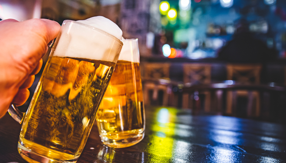 Two people doing cheers with two pints of beer over wooden table with blurred lights in background at an Appleton WI restaurant.