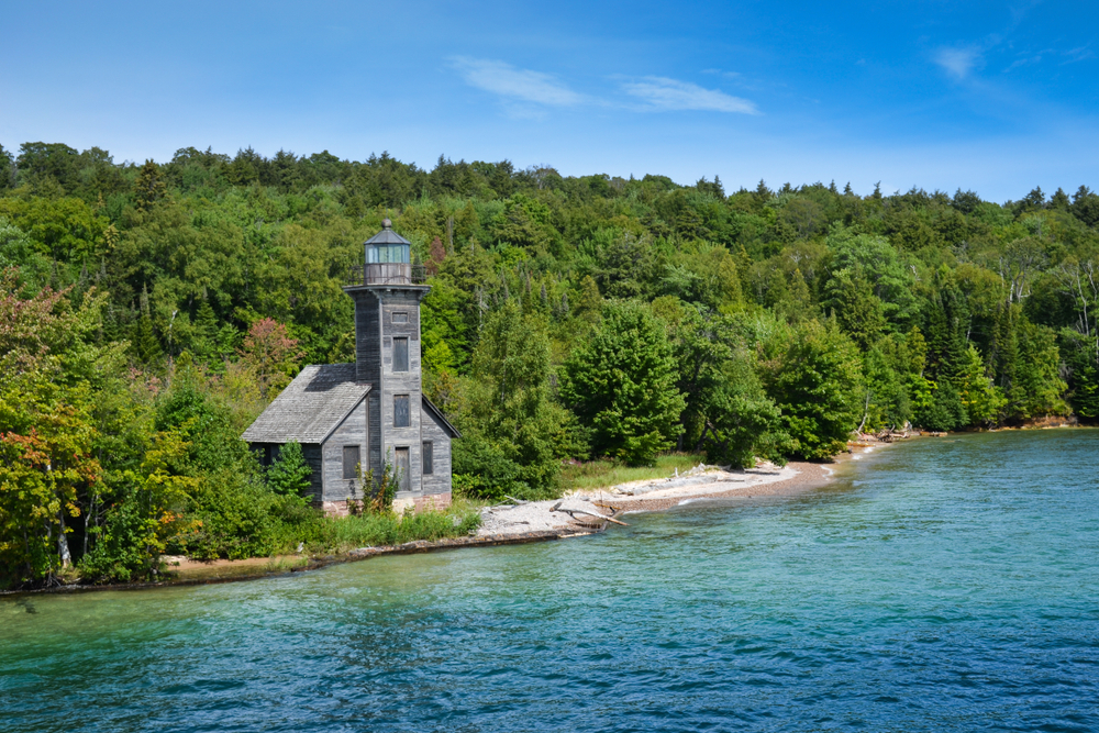 Aerial view of a wooden lighthouse surrounded by trees and overlooking the blue water of the lake.