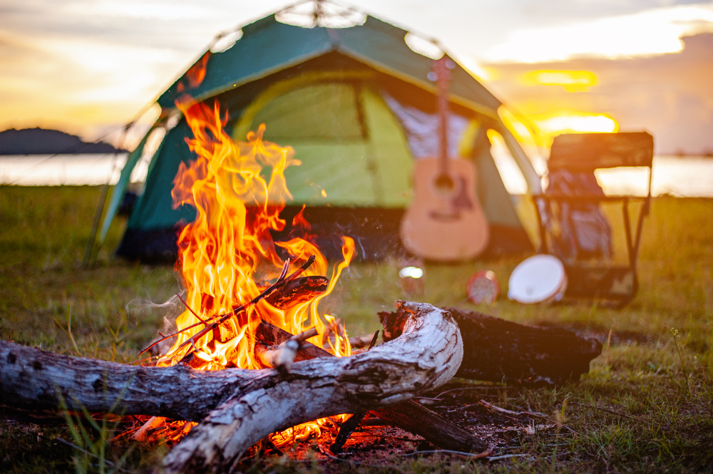 A closeup of a bonfire. In the background you can see a green tent with a guitar propped against it. There is also a camping chair with a tambourine. 