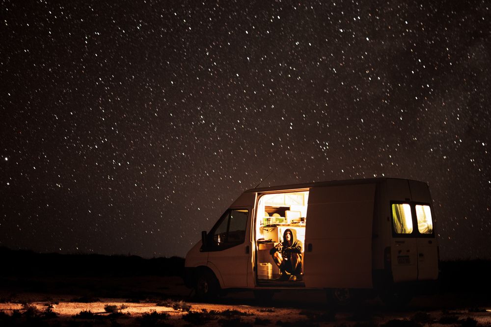 A camper van that has the door open and you can see a person sitting inside. The campervan is lit up and it is dark outside. You can see stars in the sky. 
