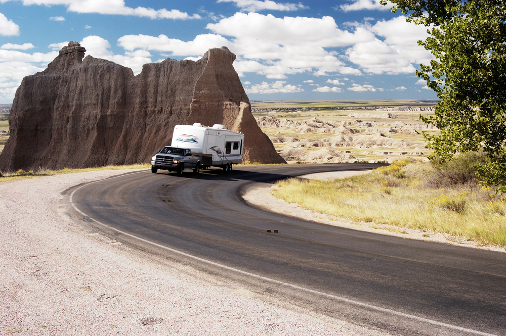 A truck pulling an RV down a winding road in the Badlands. You can see Badlands formations in the distance and it is a sunny day. 