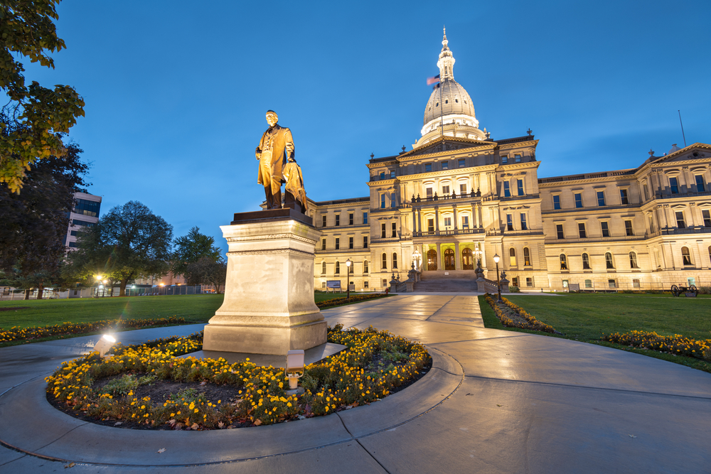 The Michigan State Capital building in Lansing. You can see a statue in front of the building and the building is lit up. 