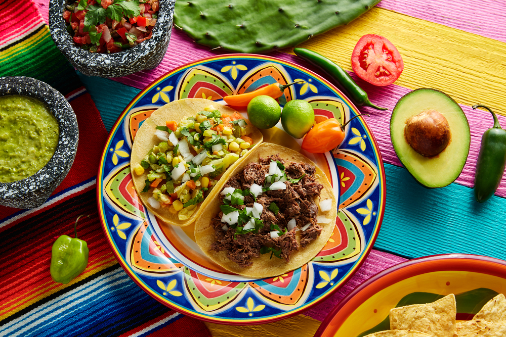 A colorful painted plate with a veggie taco and a shredded beef taco. There is also a colorful table cloth, cut vegetables, a bowl of salsa and a bowl of guacamole. It similar to what you can find in restaurants in Lansing.