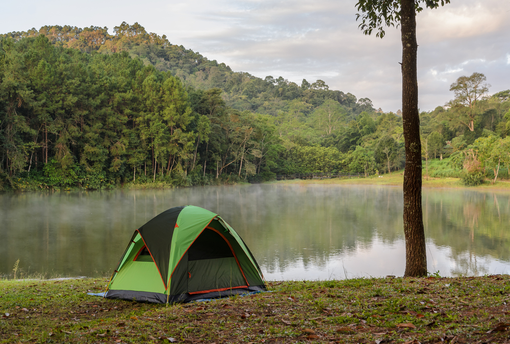 A small tent on the side of a small lake. The lake is mostly surrounded by trees, but there are some grassy patches on the lake shore. 