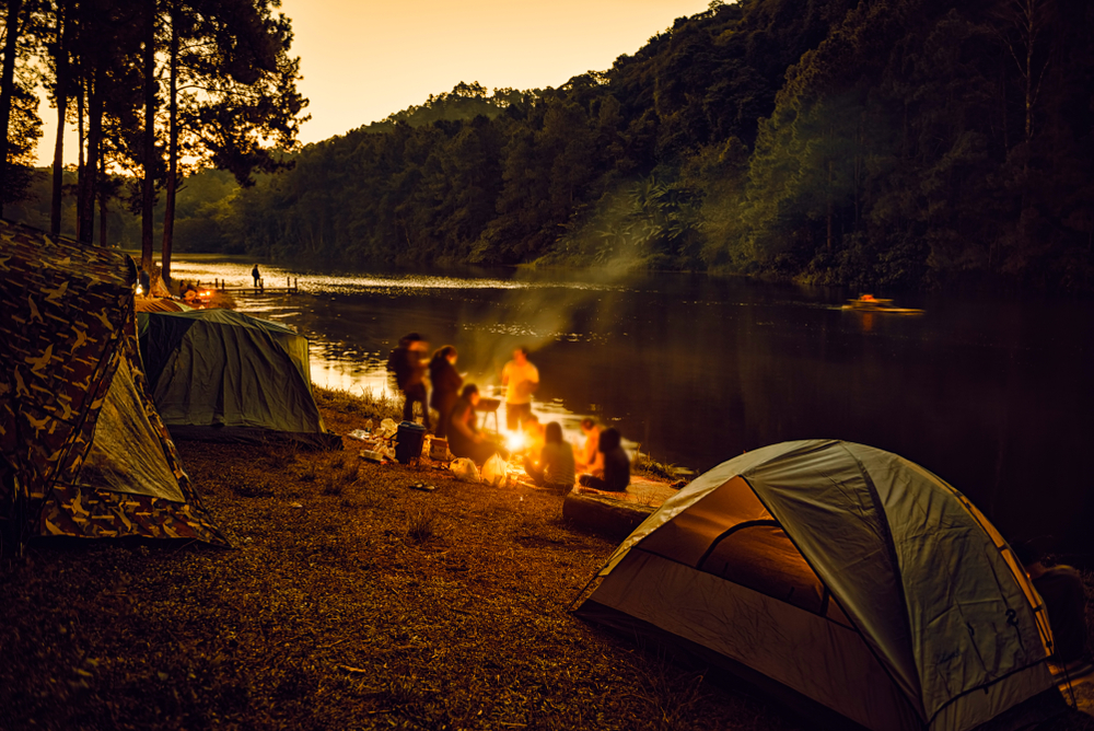 A group sitting by a campfire near tents on the shore of a river. The sun has almost completely set. 