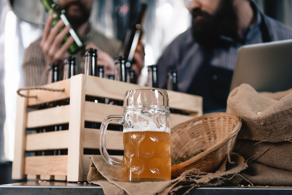 A glass of beer in front of a crate containing creaft beer two people are in the background taking beer from the crate