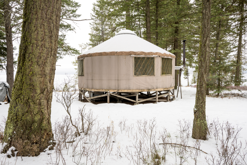 A yurt in the woods covered in snow. There are a few windows on the yurt and a metal chimney sticking o