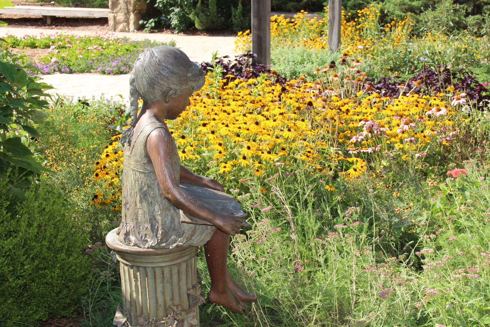 A metal statue of a girl sitting and reading a book. The statue is in a garden with yellow, pink, and purple flowers as well as tall grasses. Its one of the best things to do in Wichita KS.