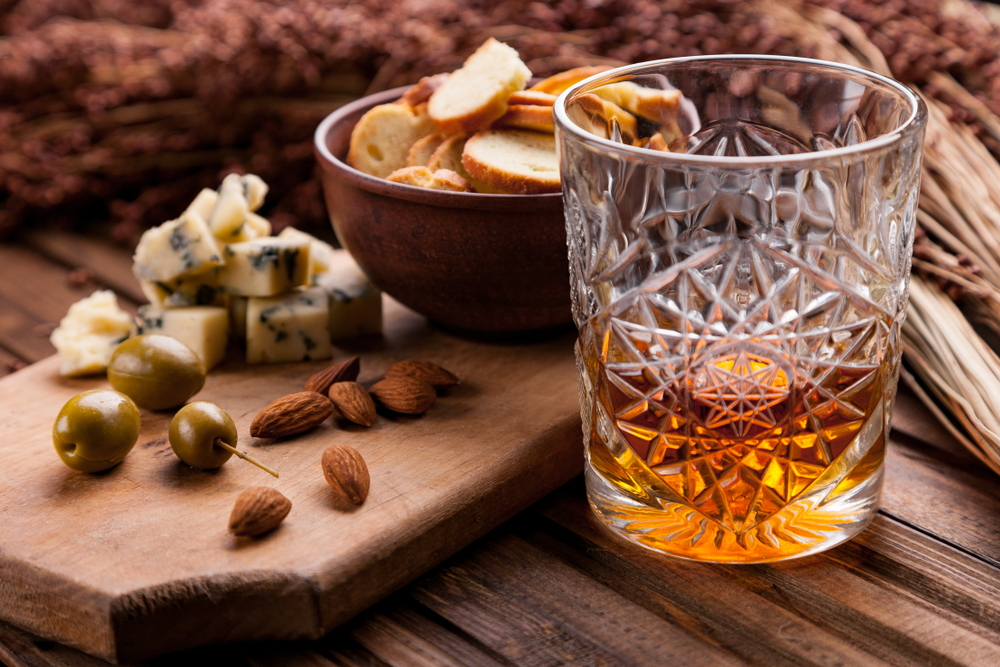 A crystal glass of bourbon next to a tray of cheese, olives, crackers, and nuts. 