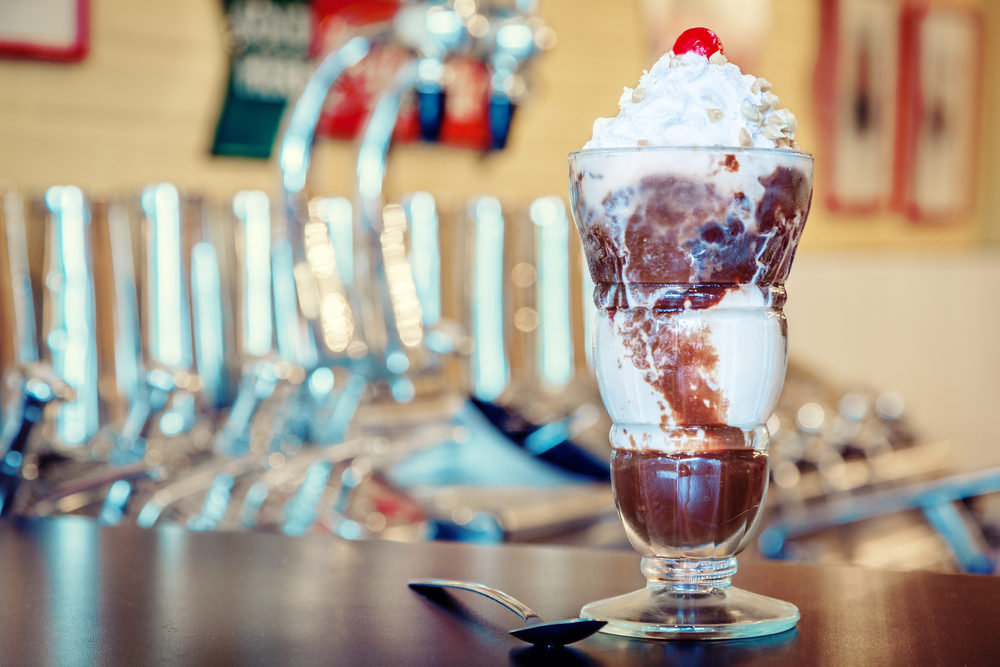 A chocolate fudge sundae in a tall clear glass with whipped cream and a cherry on top. Sitting next to the glass is a spoon. 