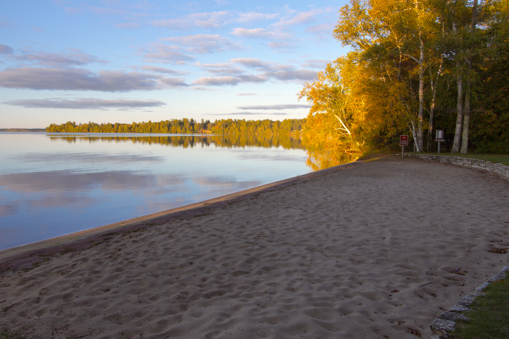 The sandy beach of a lake in Michigan. The lake is also surrounded by trees  and grassy areas. 