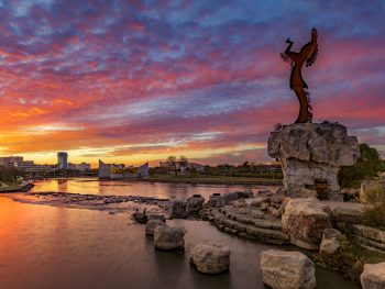The Keeper of the Plains statue looking over the Arkansas River as the sun sets over Wichita in the distance. It's one of the best things to do in Wichita.