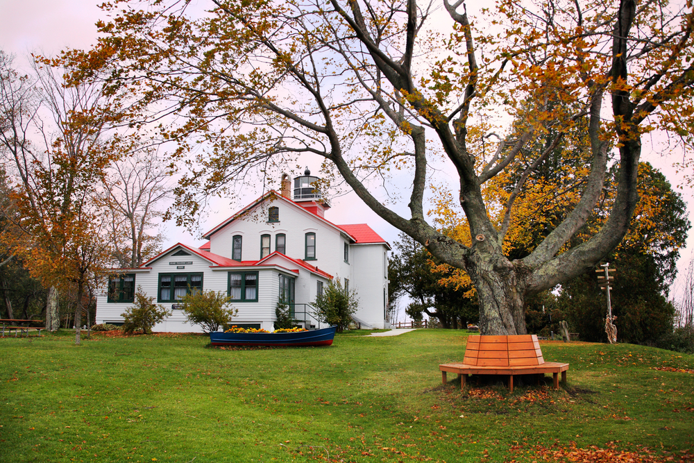 A historic white building attached to a small lighthouse. It has a red roof and is surrounded by a grassy yard with trees. 