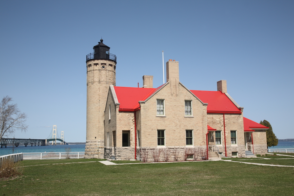 A historic building made of grey brick with a red roof. Next to it is a small grey brick lighthouse with a black top. You can see a large bridge in the water behind the building and lighthouse. 